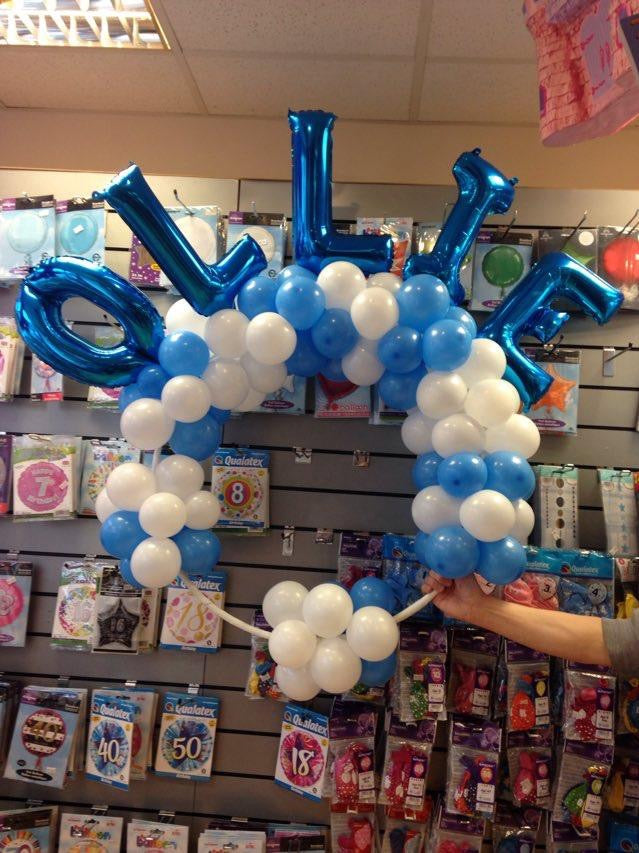 Balloons spelling the name Ollie arranged on a hoop, in blue and white. A hand holds the design in place.
